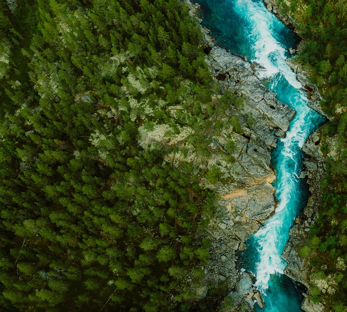 Drone high-angle photo of the turquoise-colored mountain river flowing in the pine woodland with a view of the mountain peaks in the background in Innlandet County, Norway